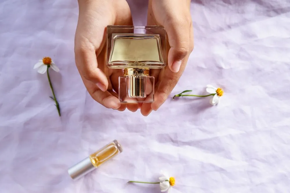 Unas manos sostienen un frasco de perfume sobre lino, entre flores blancas